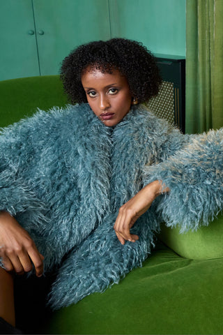 Woman in a blue fluffy coat sitting on a green couch with a lamp and green curtains in the background.